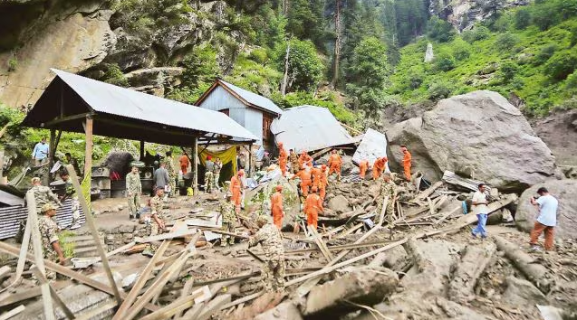 Rescue teams in orange uniforms search through debris and damaged structures after a cloudburst in Kishtwar, Jammu and Kashmir, surrounded by large boulders and forested hills.